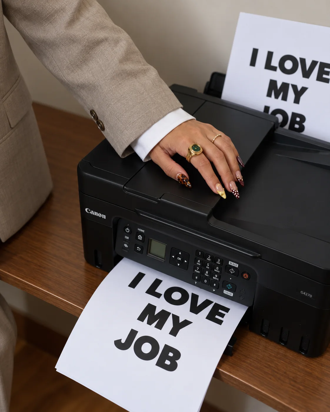 A hand with gold rings and patterned nails resting on a printer printing “I Love My Job” pages