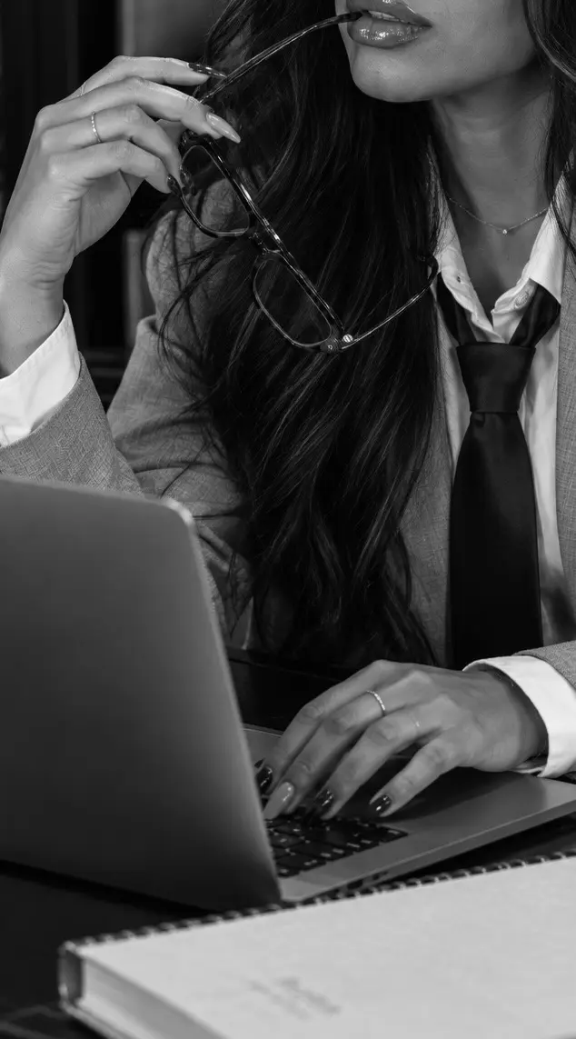 Jenica holding her glasses while typing on a laptop, in black and white